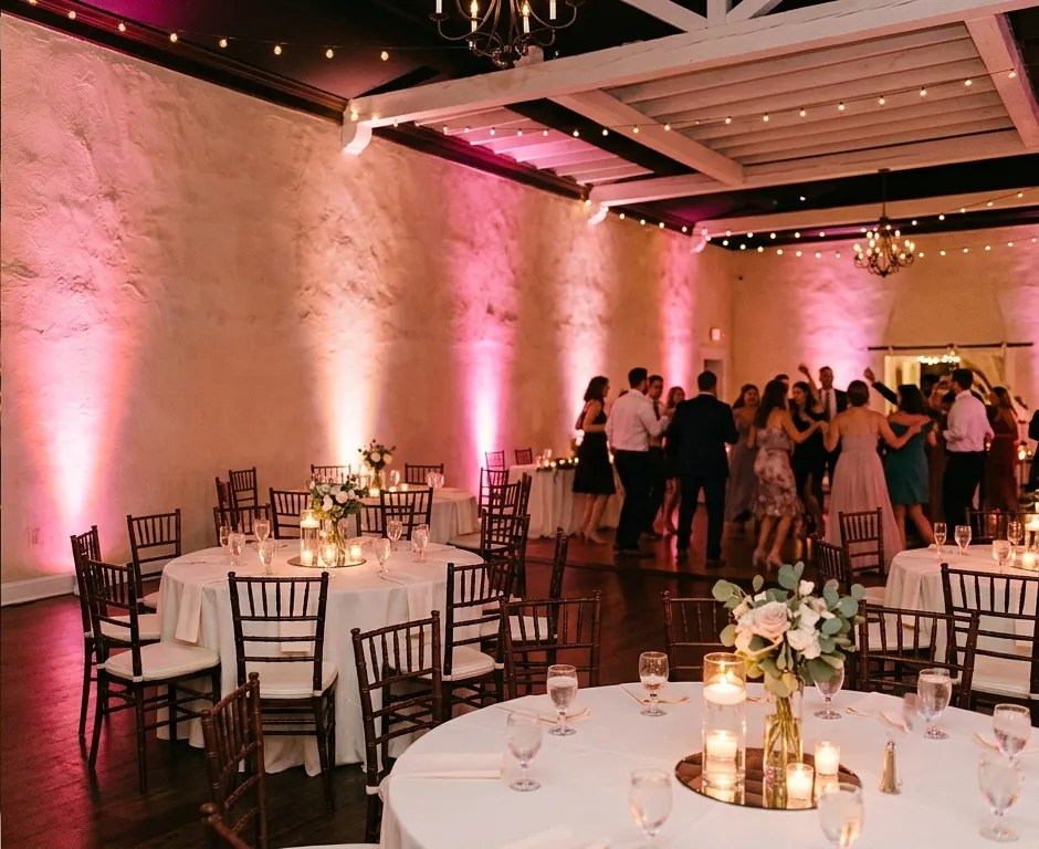 Pink uplighting rental washing textured stucco walls at an indoor wedding reception with bistro string lights and guests on the dance floor